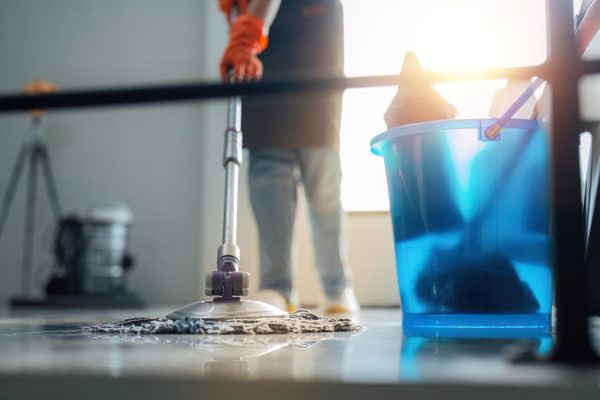 A cleaning woman is standing inside a building holding with a blue tank on the side facilities for tidying up in her hand.