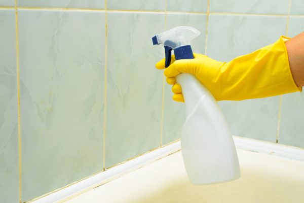 Cleaning and washing in bathroom. Close-up of female hand in yellow rubber glove using chemical spray. Side view of housewife cleaning dirty tiles in bath.