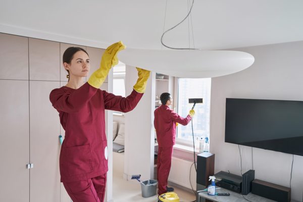 Focused woman wiping uplight bowl pendant with microfiber cloth while man steam-cleaning window